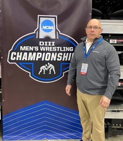 Paul Higgins stands next to banner for NCAA DIII Wrestling Championship