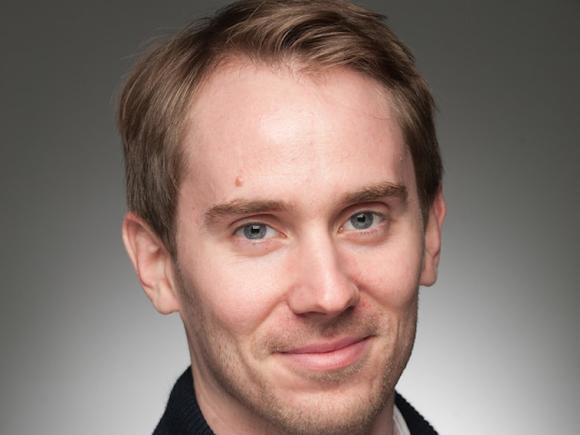 Colin Moge, class of 2016. He smiles in front of a plain, gray background, as part of his headshot. He is wearing a black sweater with a white shirt underneath. He has short, blond hair and blue eyes.
