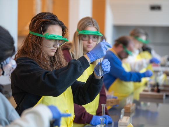 Chemistry students in a lab wearing eye protection and aprons.
