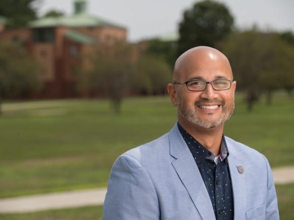 A landscape portrait of Junior Delgado, Director of the Career Center. He's wearing a navy shiny and light blue suit jacket and is standing in front of the Campus Green, which is blurred behind him.