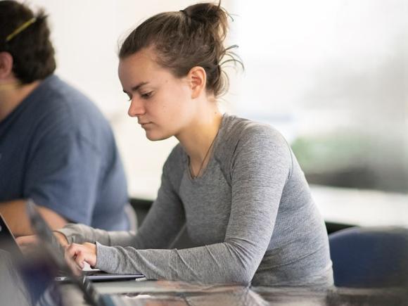 A student wearing a grey shirt, working on a laptop in class.