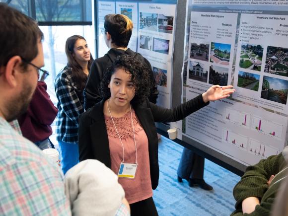 Student presenting research pointing at a poster chatting with other students.
