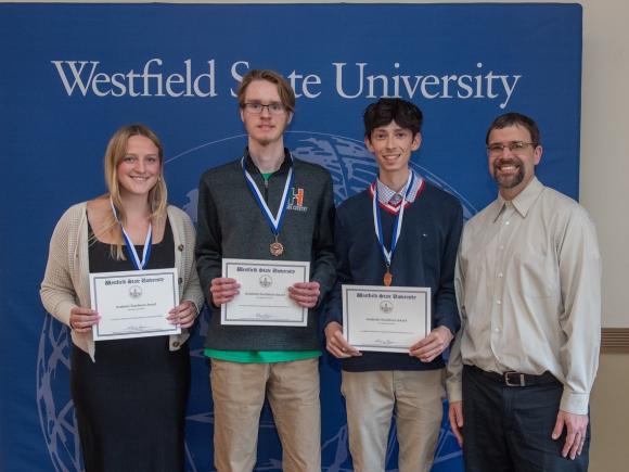 Three students smiling while holding academic award certificates at an academic awards ceremony.