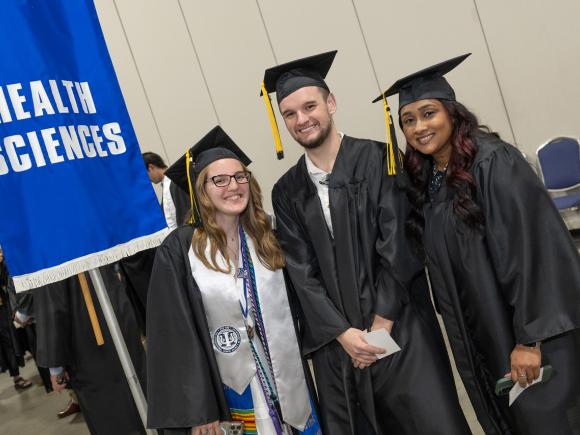 Three Health Sciences students smiling in graduation attire at commencement, standing in front of a Health Sciences flag.