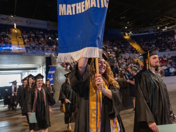 Math students wearing caps and gowns at commencement, proudly holding the Mathematics Department flag.