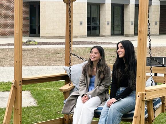 Two students smiling together on a wooden swing. 