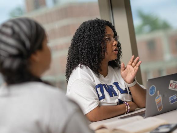 Student speaking with classmates while seated in front of a laptop.