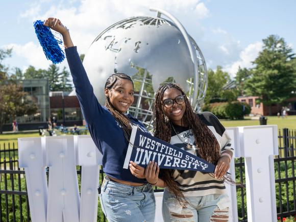 Two female students stand holding a blue Westfield State University pennant and pom pom in front of the Globe statue
