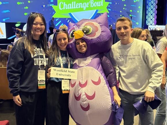 Three students stand with a fourth student wearing an owl costume in front of the logo for the PA Medical challenge