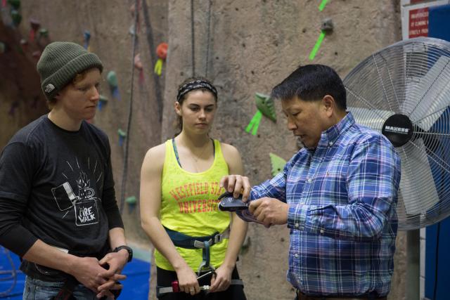 Two movement science students speaking with instructor in front of rock climbing wall.