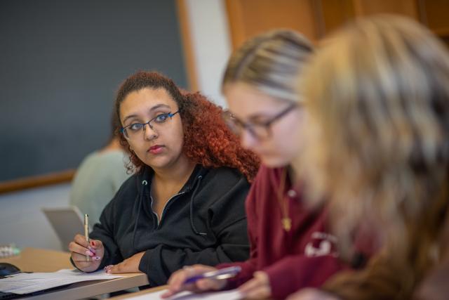 Three students in history class writing with one student looking up with long red hair and black shirt.
