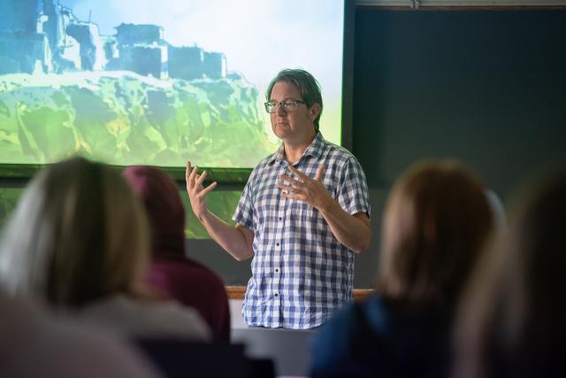 History professor Dr. Aieta wearing blue and white plaid shirt standing in front of projection screen in classroom.