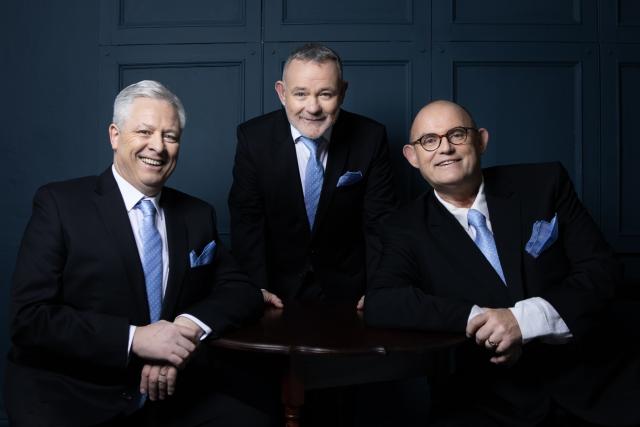 Three members of the Irish Tenors smiling wearing black suits with blue ties.