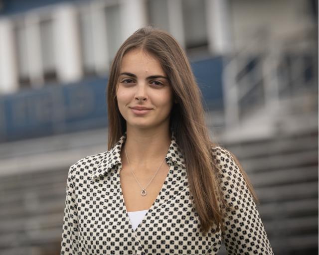 Student smiling while standing on an athletics field, with sports facilities visible in the background.