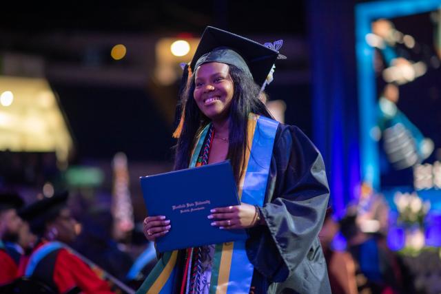 Student smiling at commencement ceremony wearing cap and gown holding diploma.