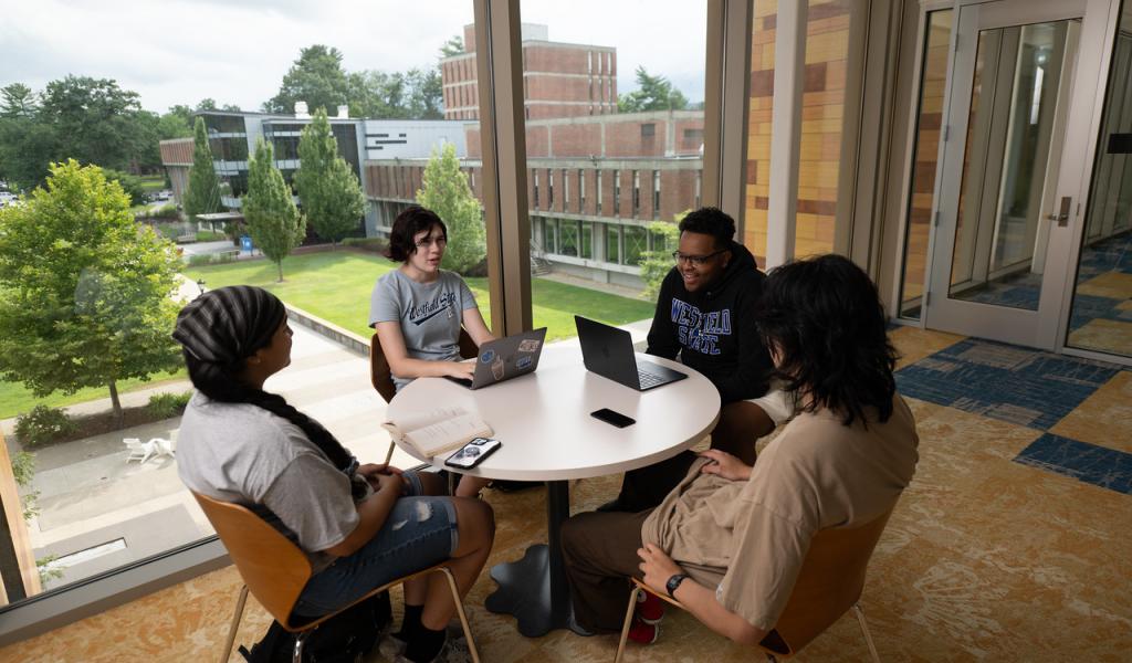 Group of students sitting around a round table chatting with laptops.