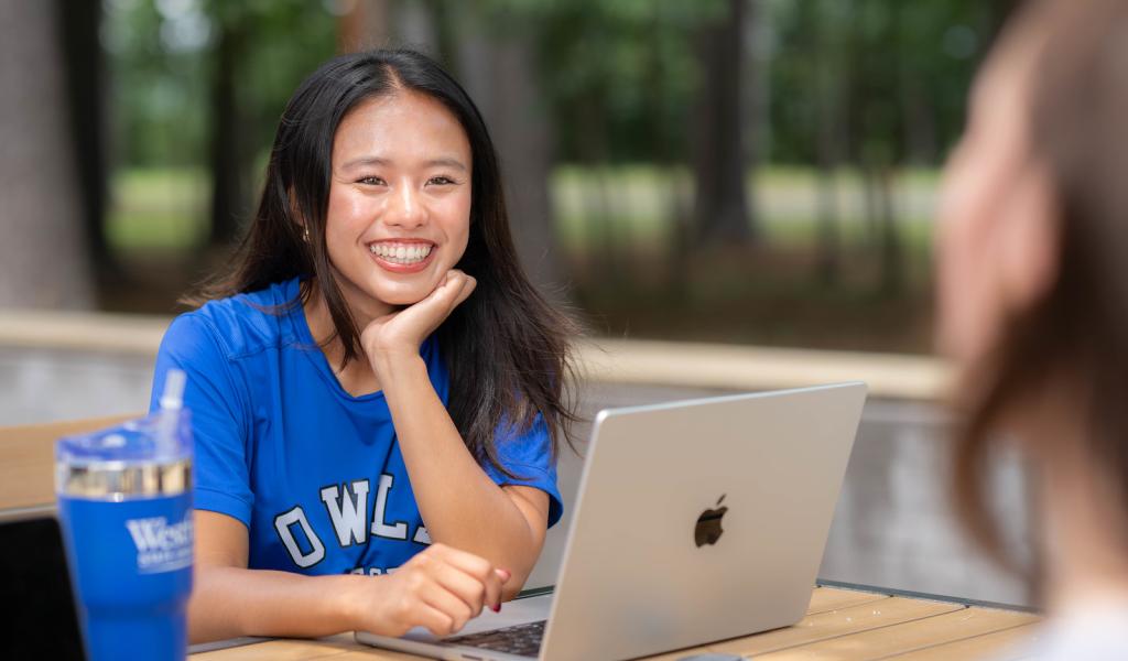 Student studying outside with a laptop while talking to another student.