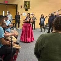 Woman sings to audience with row of drummers and dancers